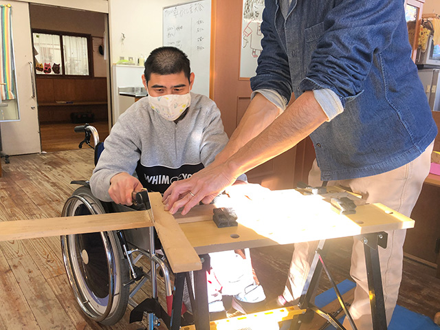 長い木材を切る車椅子に乗った男性と、補助する男性