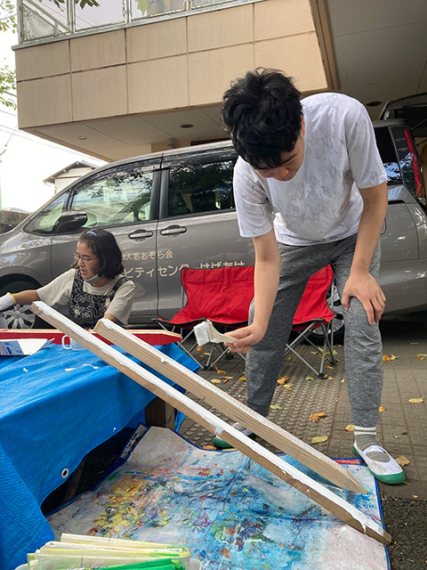 斜めにセットした2本の角材に、刷毛で白いペンキを塗る男性
