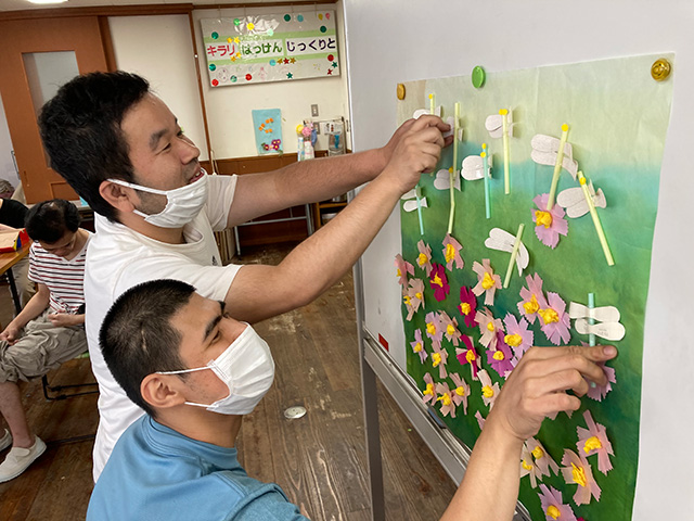 壁に貼った大きな上に「トンボ」「コスモス」の作品を貼る男性2人